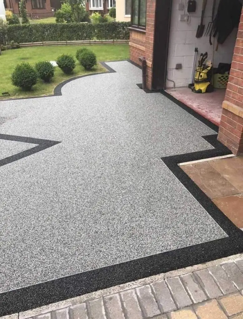 Smooth, light gray driveway with a black border, featuring a wavy edge design, leading to a brick garage with garden tools inside and neatly trimmed bushes on the adjacent green lawn.