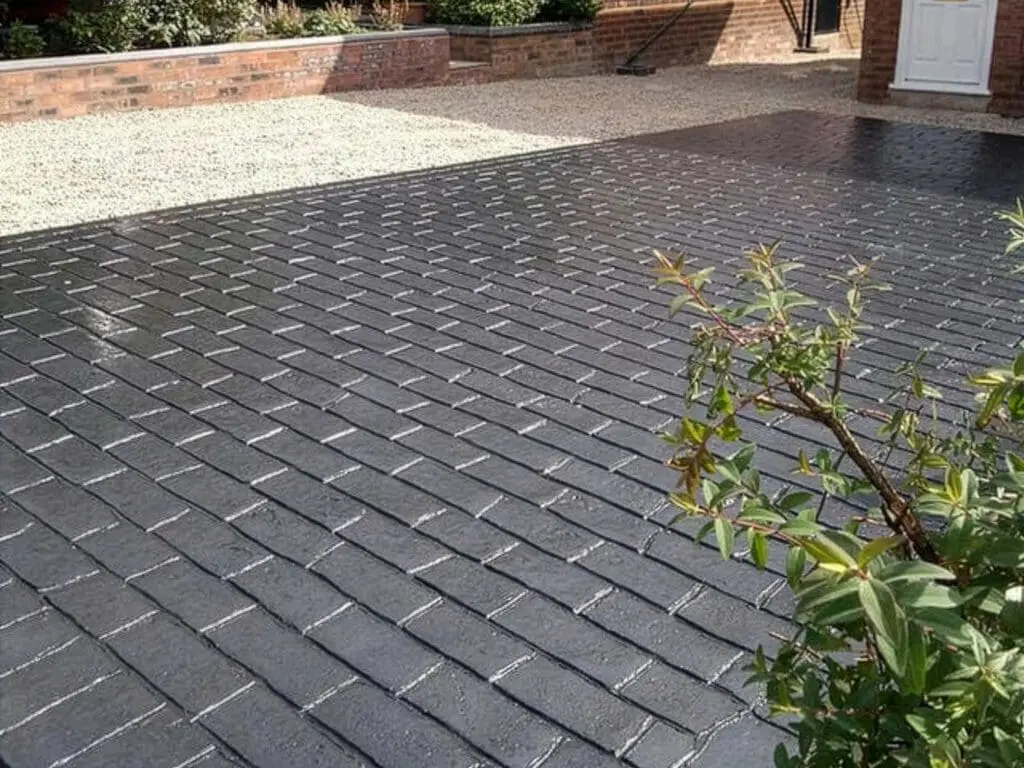 A driveway with a black brick-patterned surface is in the foreground, bordered by a gravel area and a low brick wall. Some greenery is visible on the right side of the image.
