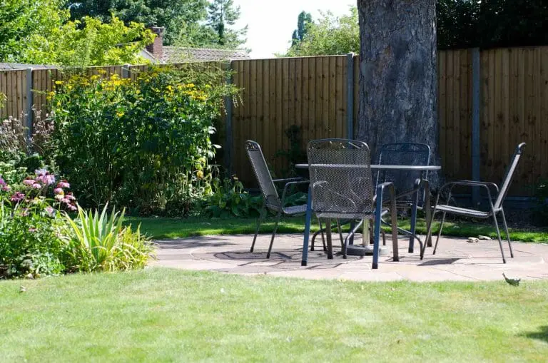 A metal patio table with four chairs sits on a stone patio in a sunny backyard, surrounded by green grass, plants, and a wooden fence.