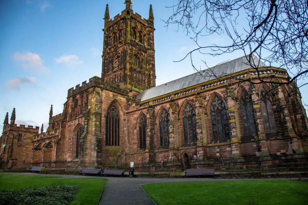 A large, historic stone church with stained glass windows and a tall central tower, surrounded by green lawns and benches, under a blue sky with some clouds.