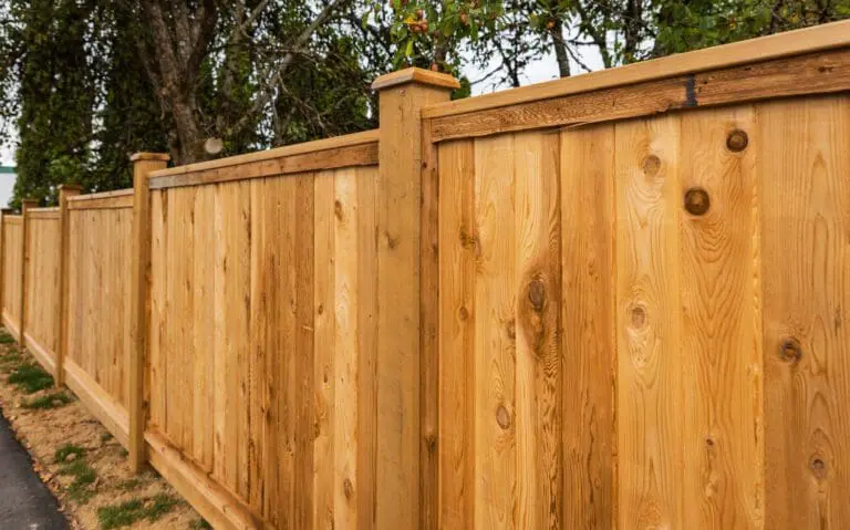 A close-up view of a tall wooden fence with vertical planks and sturdy posts runs alongside a grassy area and trees in the background.