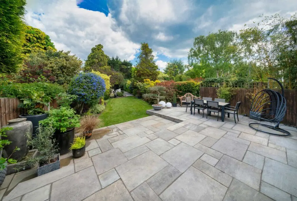 A spacious backyard with a stone patio, outdoor dining set, hanging chair, and lush green garden surrounded by trees and shrubs under a partly cloudy sky.