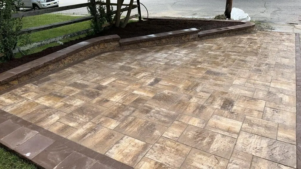 A newly installed patio with a pattern of tan and brown square and rectangular pavers, bordered by brown edging, next to a raised garden bed and a wooden fence.