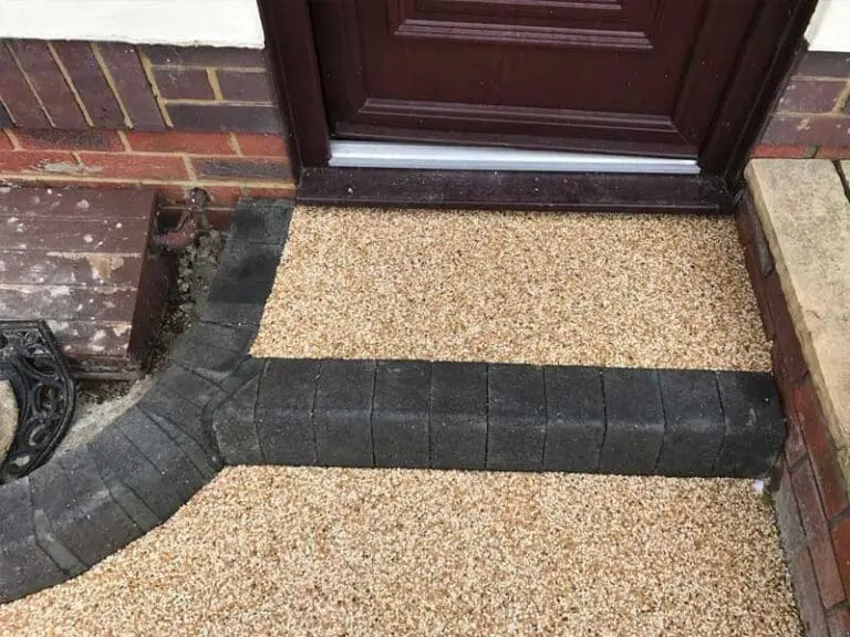 A resin-bound gravel pathway with dark brick edging leads to a dark wooden door. The bricks form a curved border, and the path meets a red brick wall and stone step.