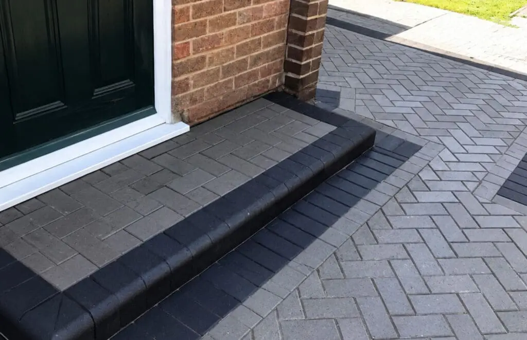Gray and black brick pavers form a step leading to a dark green door, with herringbone-patterned paving outside a brick building. Sunlight casts shadows on the walkway.