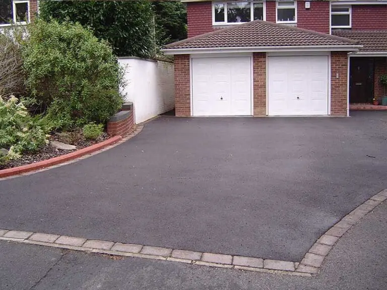 A house with a large, clean asphalt driveway leading to a double garage with two white doors. There are plants and shrubs along the left side of the driveway.