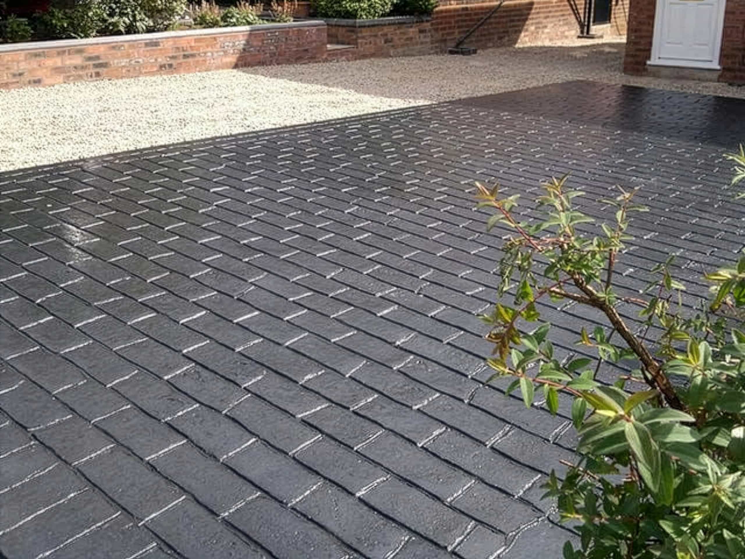 A driveway with a black brick-patterned surface is in the foreground, bordered by a gravel area and a low brick wall. Some greenery is visible on the right side of the image.