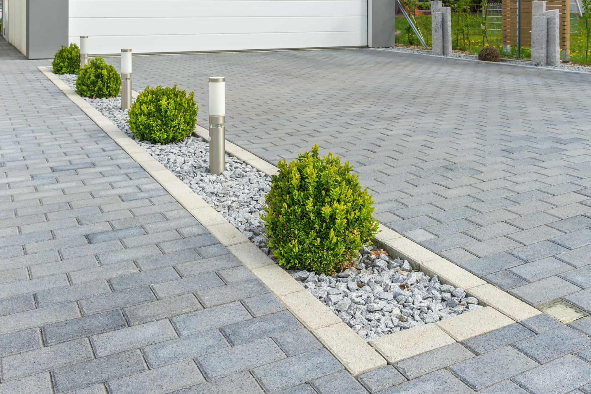 A paved driveway features a central strip with small green bushes, gray rocks, and modern outdoor lights, bordered by light-colored stone edges.