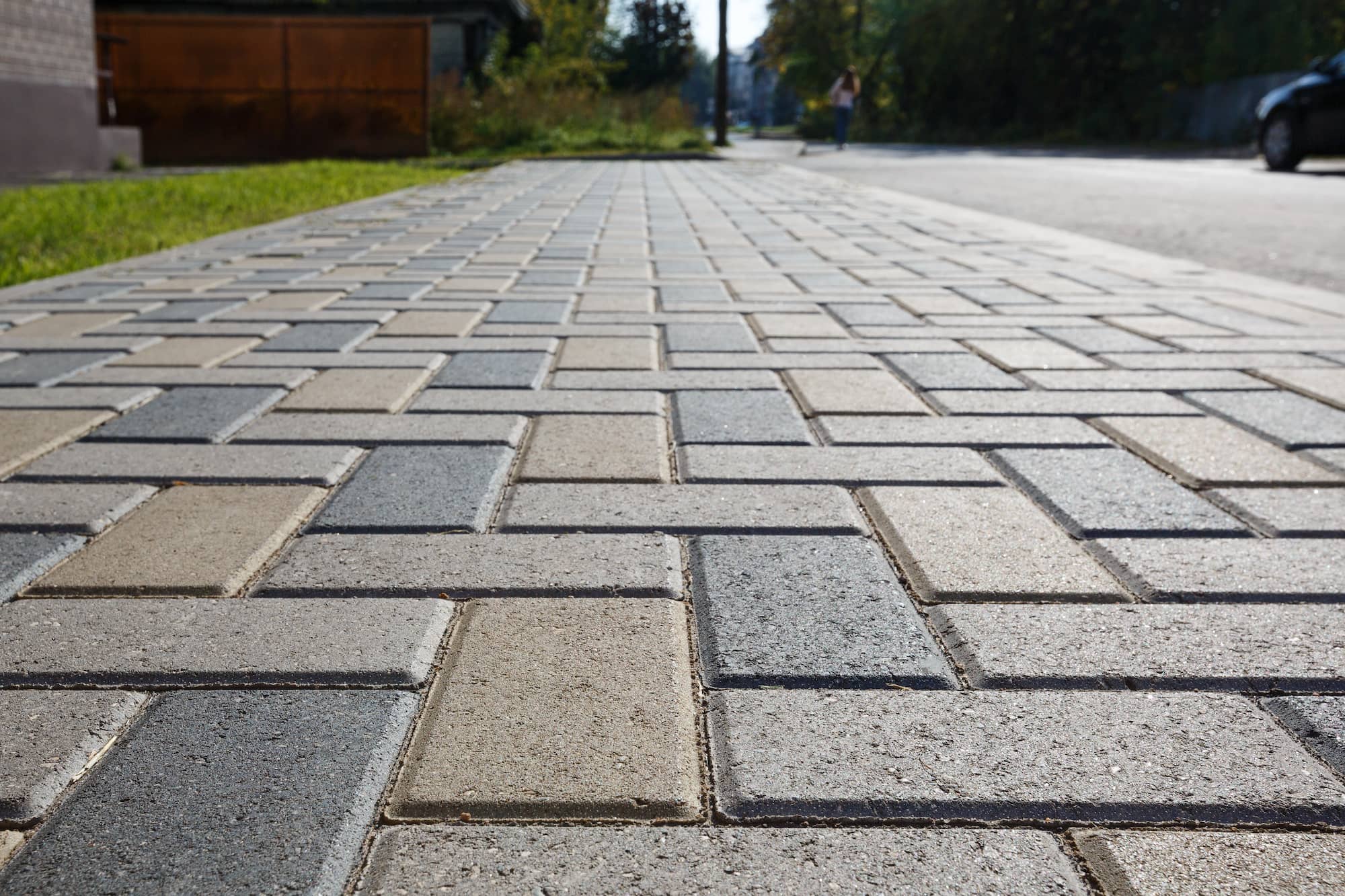 A close-up view of a paved sidewalk with rectangular bricks in a herringbone pattern, leading toward a street with trees, a fence, and a person in the distance.