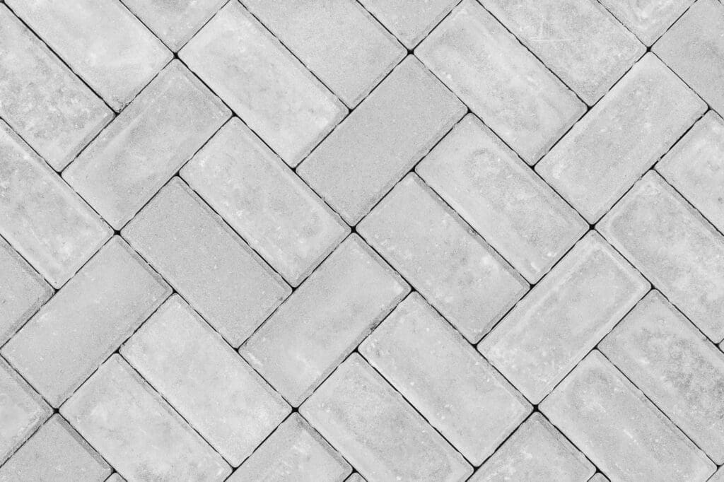 Gray rectangular paving stones arranged in a herringbone pattern, creating a diagonal, interlocking grid design. The surface appears clean and evenly spaced, emphasizing the geometric layout.