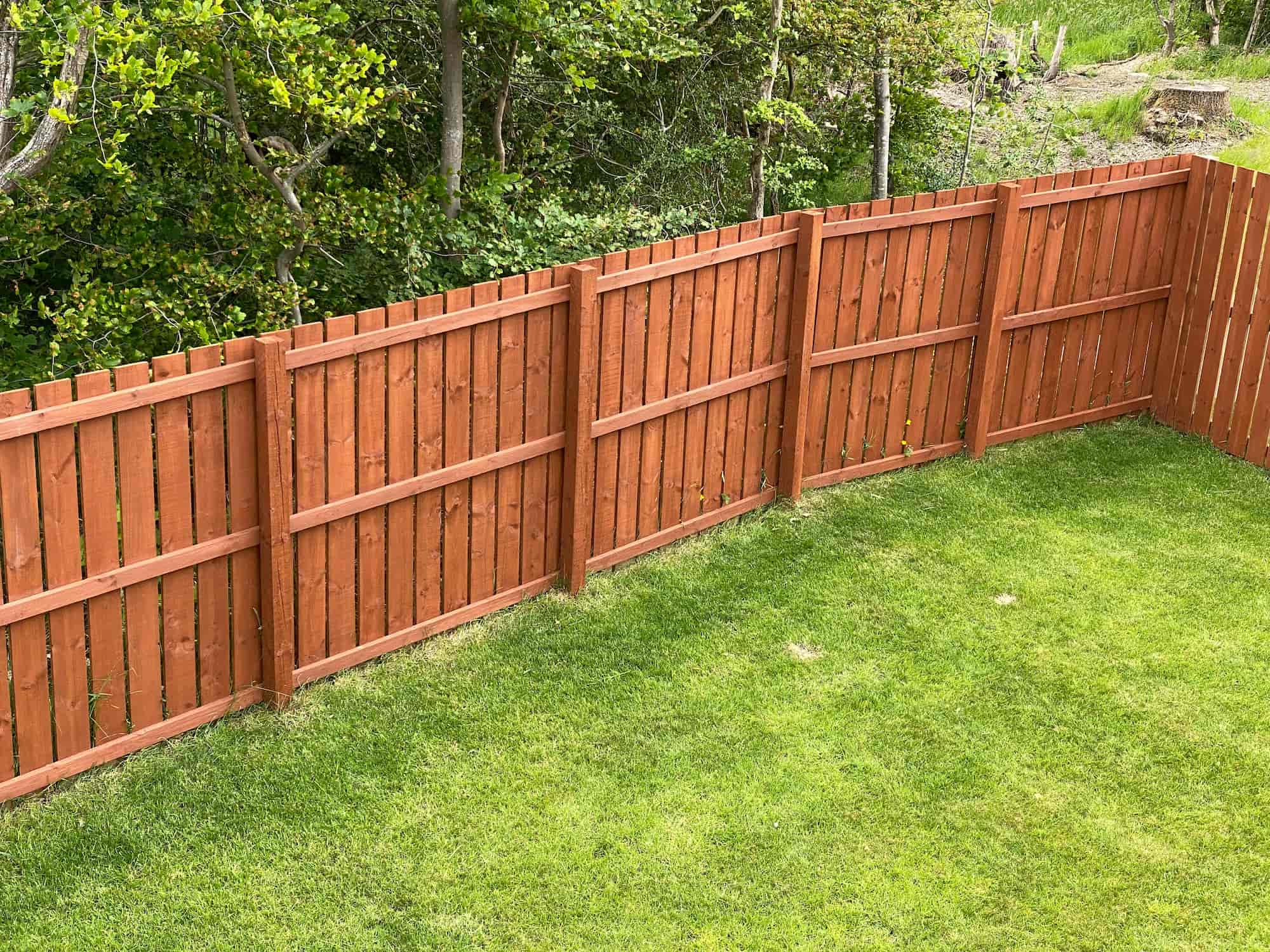 A wooden fence with vertical panels stands on green grass, with leafy trees in the background. The fence is stained reddish-brown and forms an angled corner.