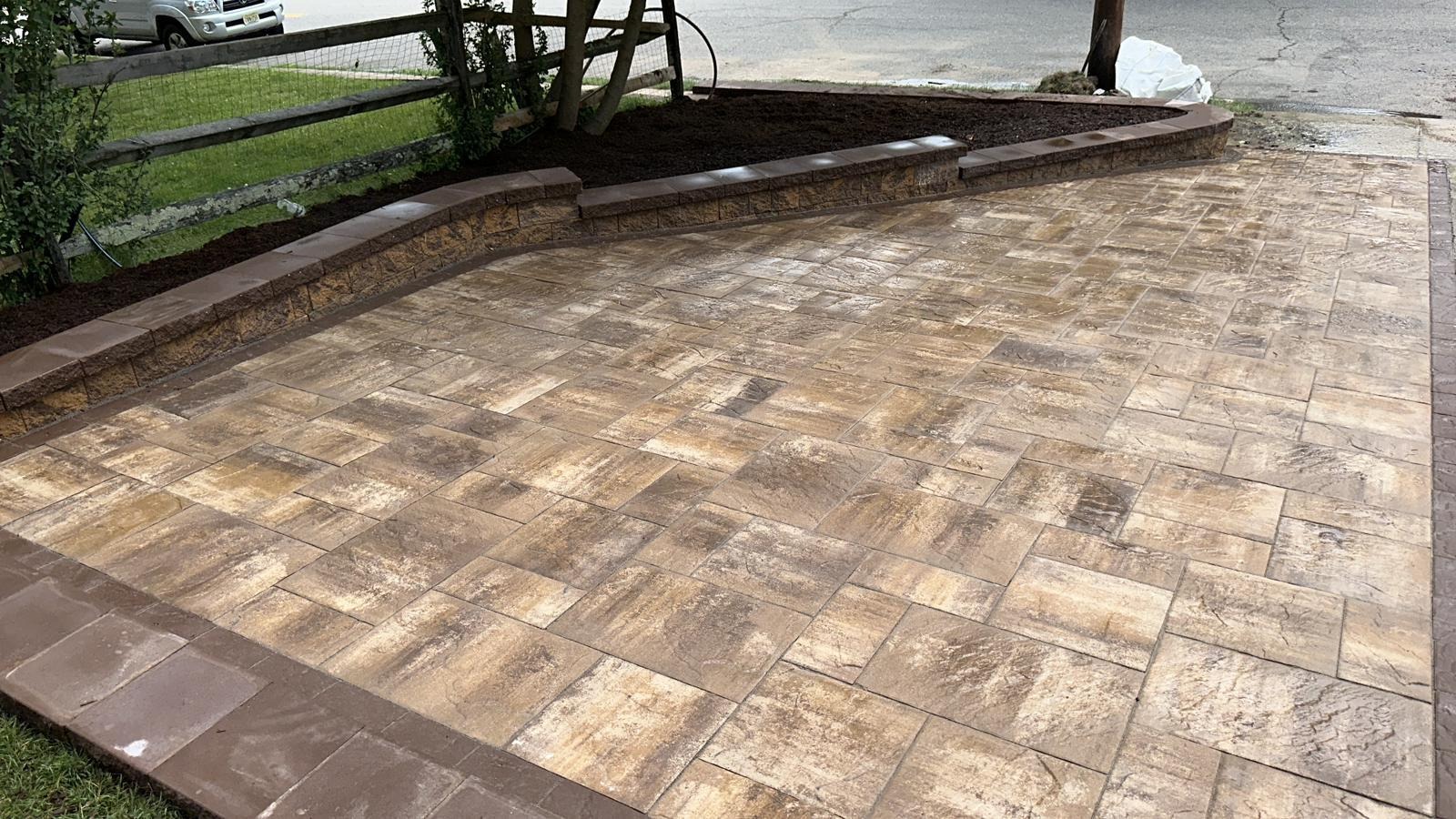 A newly installed patio with a pattern of tan and brown square and rectangular pavers, bordered by brown edging, next to a raised garden bed and a wooden fence.
