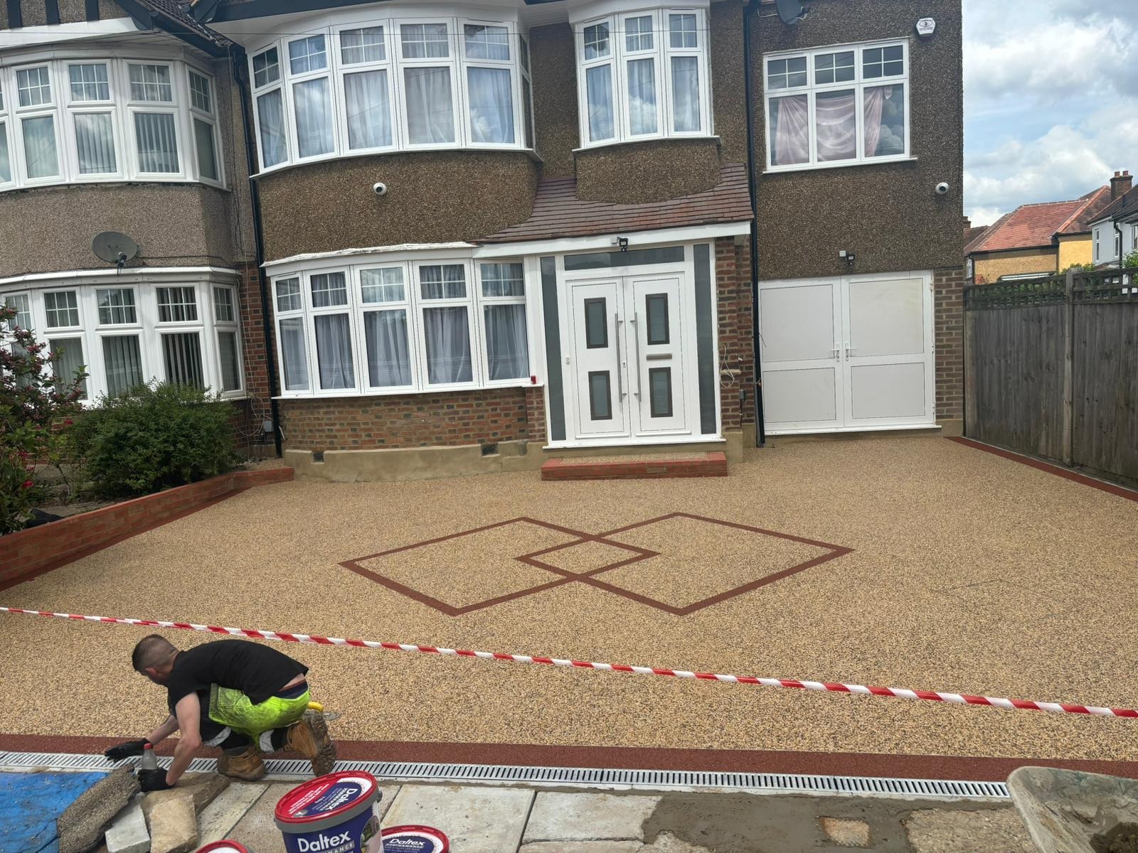 A person works on the edge of a newly finished, patterned driveway in front of a brown house with white windows and doors. Red tape separates the work area from the driveway.