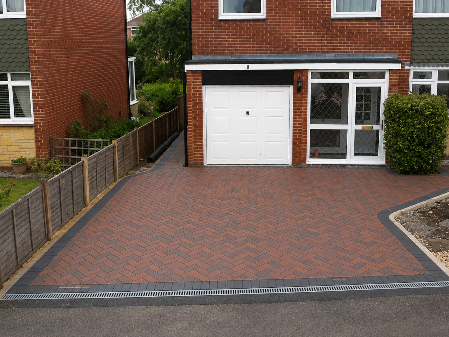 A herringbone-patterned brick driveway leads to a white garage door on a red brick house with a glass porch and a garden area at the side.