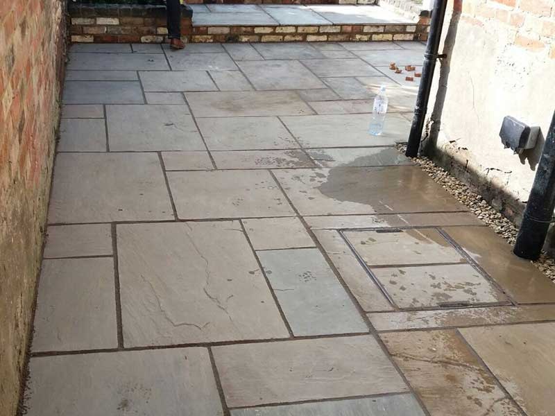 A freshly laid stone patio with rectangular tiles in a geometric pattern. There is a wet patch, some scattered pebbles, a plastic bottle, and brick steps leading upwards at the end.