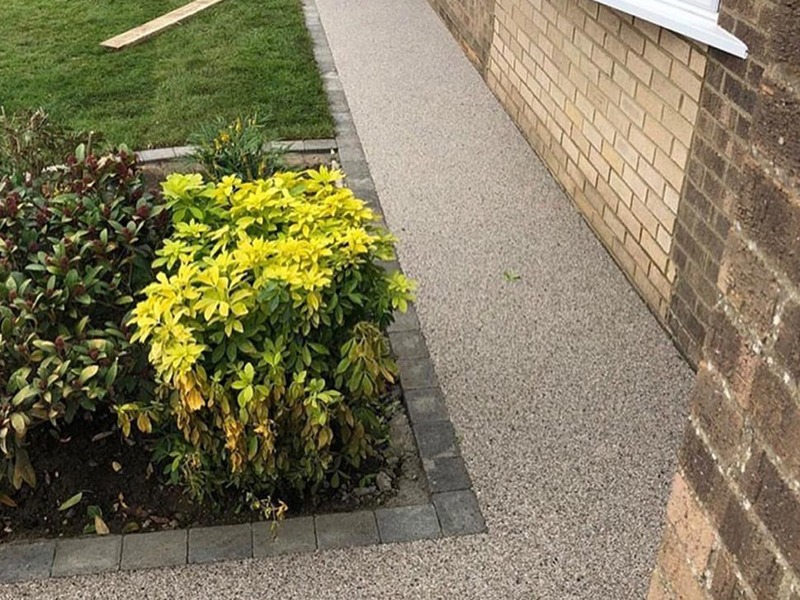 A small garden with green and yellow shrubs is bordered by a paved path next to a brick house. A strip of grass is visible in the background.