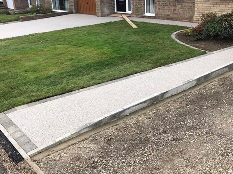 A new sidewalk runs beside a green lawn in front of a brick house. The sidewalk is elevated, with no ramp connecting it to the street, making it inaccessible for wheelchairs or strollers.