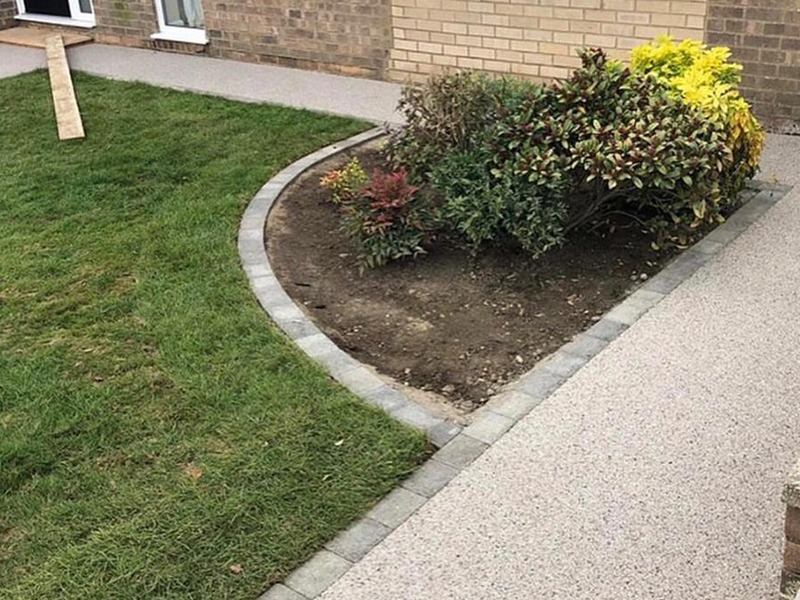 Curved garden bed with shrubs surrounded by a gray stone border, next to a grass lawn and gravel pathways, in front of a brick house.
