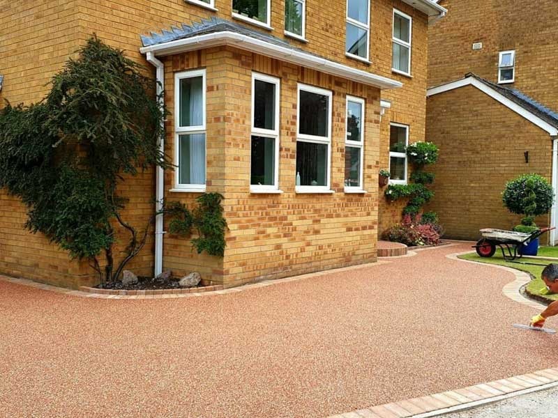 A brick house with white-framed windows, a tidy garden, and a newly installed resin driveway. A person is seen working near the edge of the driveway with a wheelbarrow in the background.