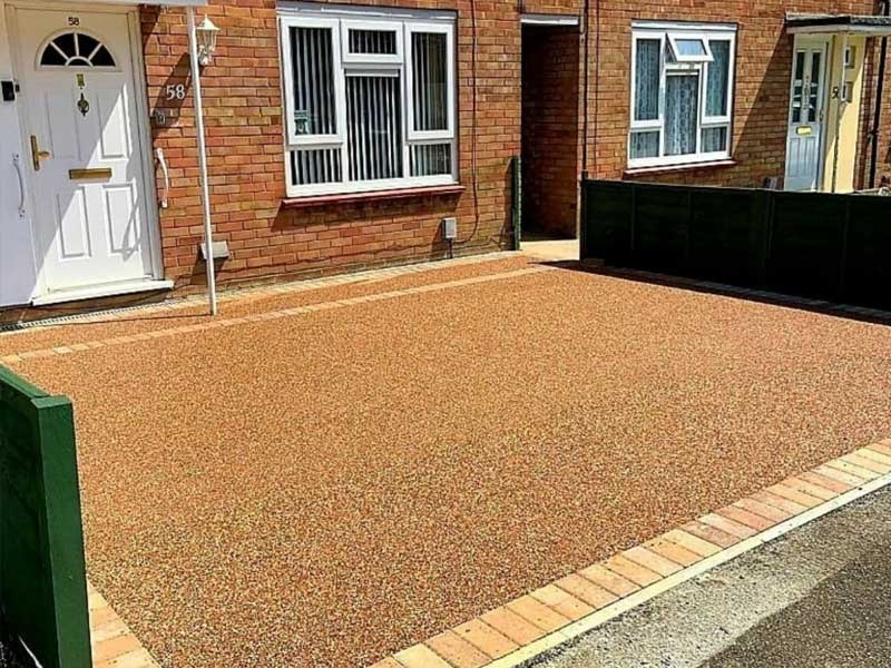 A paved driveway made to look like a gravel surface sits in front of a brick house with a white door and windows, bordered by dark green fences.