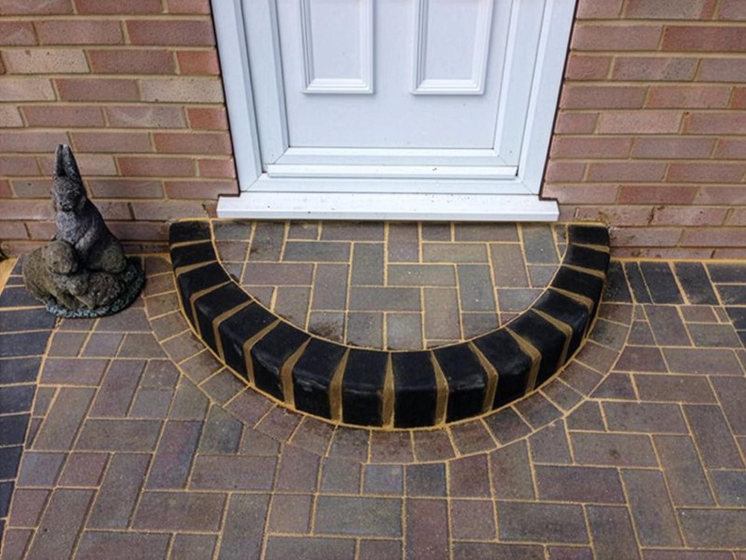 A curved brick step in front of a white door, with a stone statue resembling a small animal on the left. The bricks are arranged in a herringbone pattern.