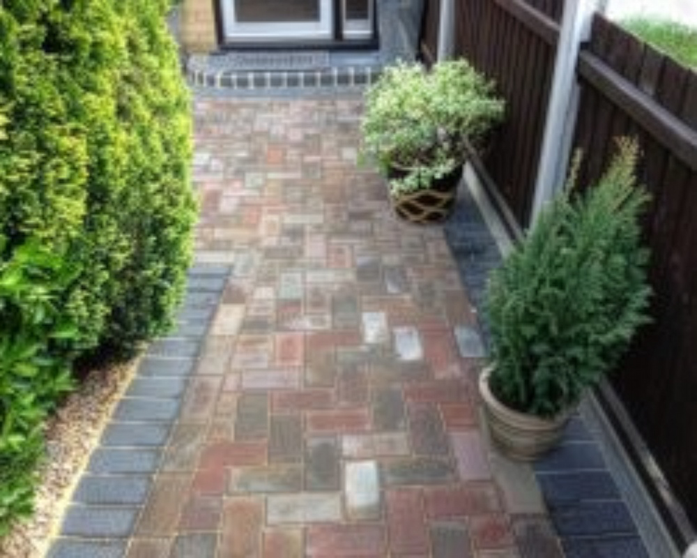 A narrow brick pathway lined with a black brick border, flanked by green shrubs on the left and two potted plants on the right, leading to a door.