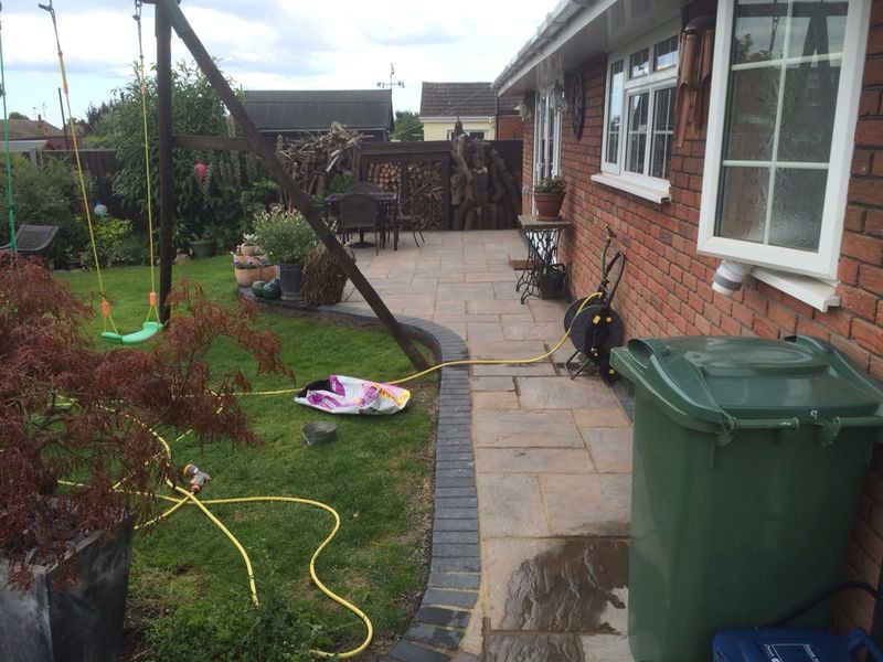 A backyard with a green swing, patio, garden hose, green bin, and potted plants. There is a bag of compost on the ground and logs stacked by a wooden fence near the patio area.