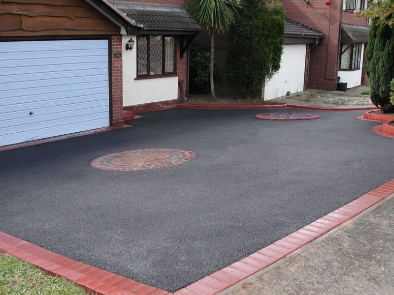 A black asphalt driveway with red brick edging and two circular brick patterns is in front of a house with a white garage door and brown trim.