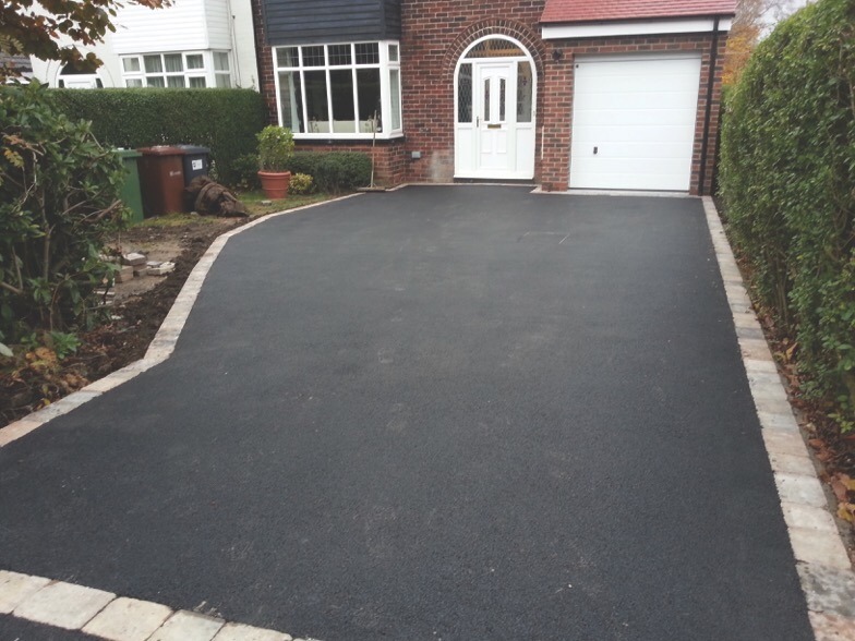 Freshly paved driveway with stone edging leading to a house with a white front door and attached garage; hedges and plants line the sides.
